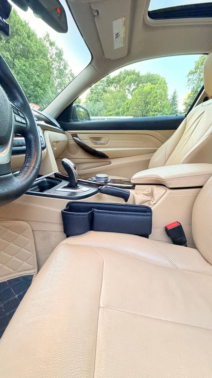 DriveTidy organizer installed in the seat gap of a car with beige leather interior, shown from the front with the dashboard and steering wheel visible.