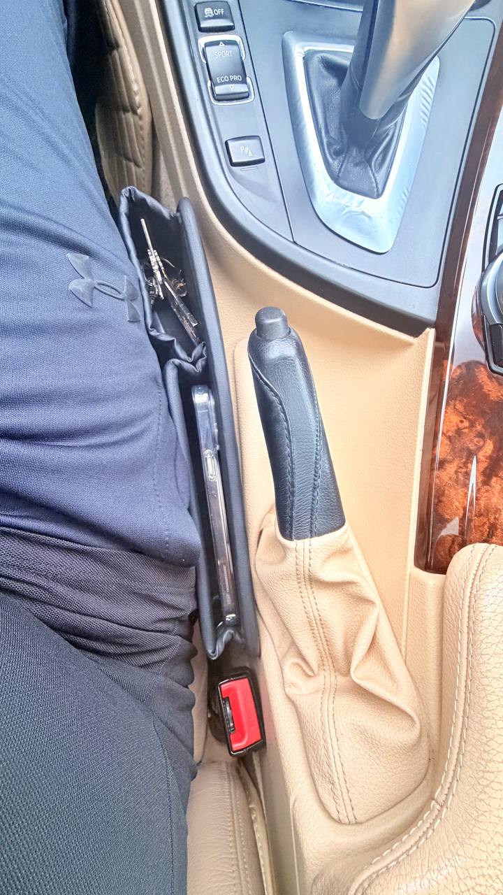 DriveTidy organizer in everyday use, holding keys and a phone in its pockets while seated in the gap next to the handbrake and beige interior.